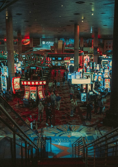 A bustling casino floor filled with groups of slot machines and people walking around. The atmosphere is vibrant with neon lights and illuminated signs. The interior has a ceiling supported by large pillars and there are stairs leading down to the gaming area.